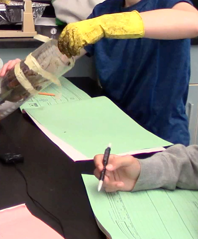 Student wearing a glove putting dirt into a bottle in a science class.